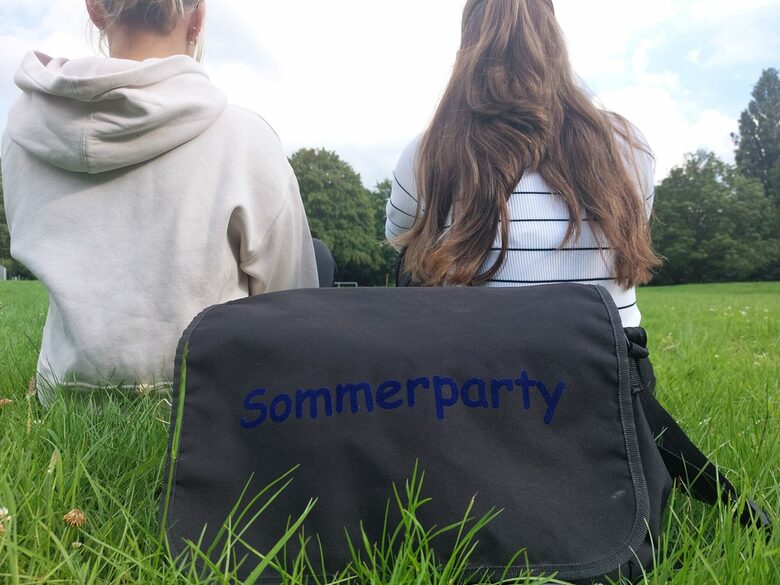 Tasche mit der Aufschrift Sommerparty liegt auf einer Wiese und zwei Mädchen sitzen mit dem Rücken zur Kamera gewandt dahinter